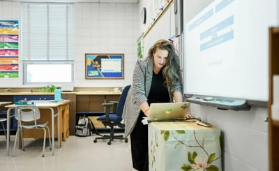 A female teacher using a laptop while standing at an interactive whiteboard in a bright, modern K-12 classroom. Featured Image