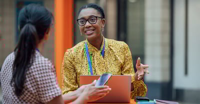 Business women sitting at a desk with a laptop talking with someone holding their phone Featured Image