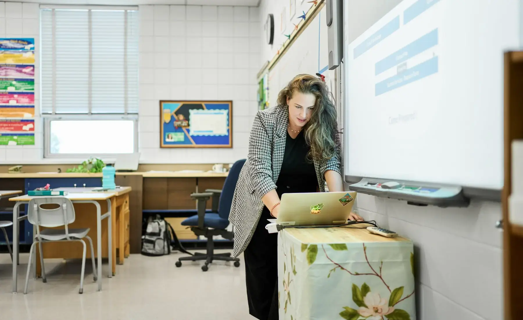A female teacher using a laptop while standing at an interactive whiteboard in a bright, modern K-12 classroom. Featured Image