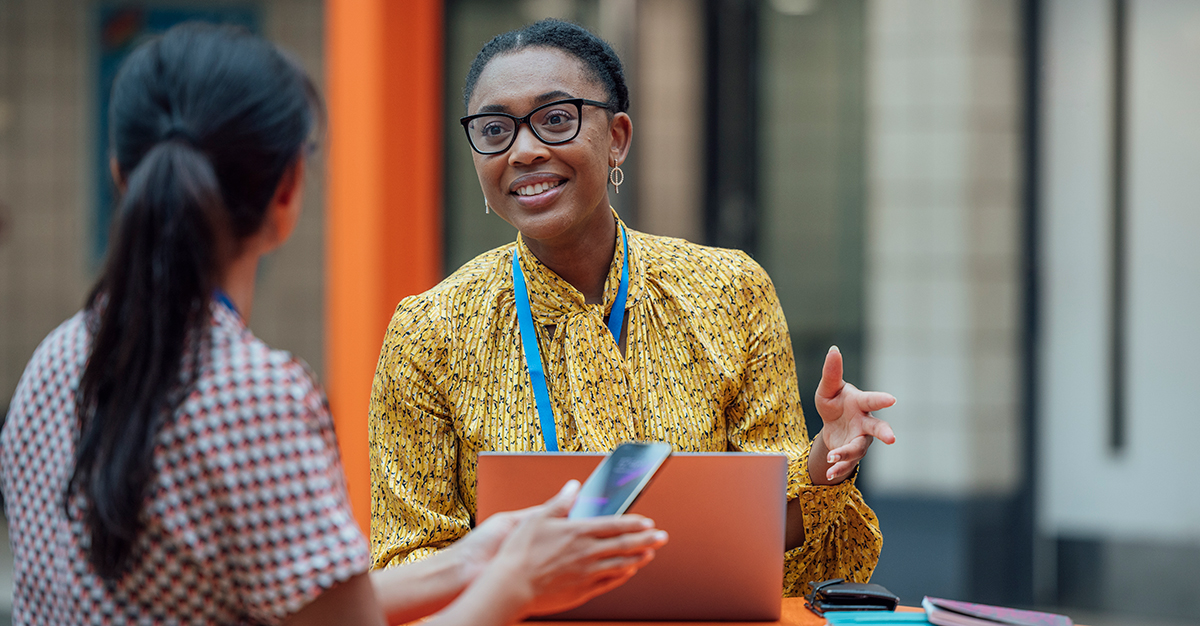 Business women sitting at a desk with a laptop talking with someone holding their phone Featured Image